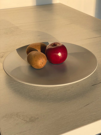 Three fruits, a red apple and two brown kiwis, are displayed on a round gray plate on a light wooden...