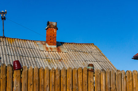 Old brick chimney on slate roof of old house behind wooden fence against blue sky.の写真素材