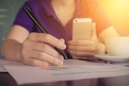 close up hand of business women holding pen pointing on paper garph chart  and using smart phone on wooden desk indoor office with morning light. vintage filter effect.の写真素材
