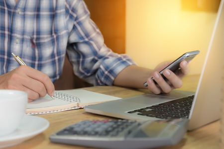 business man using smart phone and write on notebook and calculator and laptop on wooden desk outdoor office with morning light.の写真素材