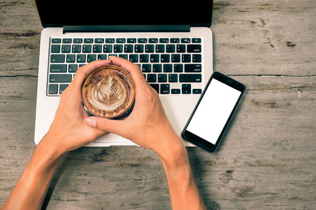 hands holding cup of coffee and laptop and smart phone on wooden background with vintage filter effect, top view.の写真素材