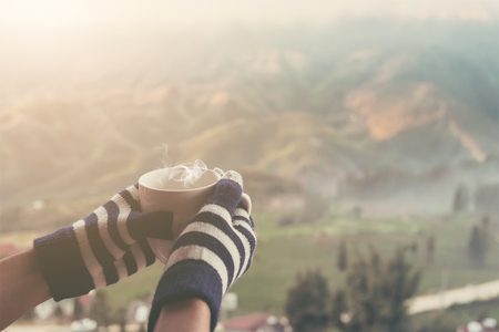 hands holding hot cup of coffee or tea in morning sunlight with beautiful mountain with fog and cloudy background. vintage filterの写真素材