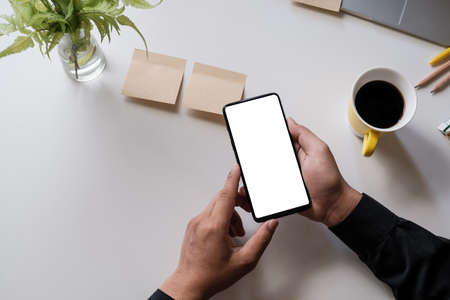 Close up of a man holding smartphone with blank screen mobile on wooden desk in home office, top...