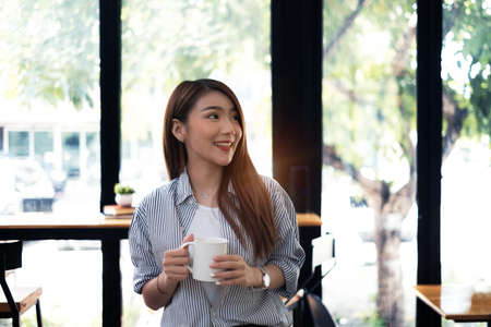 Young beautiful woman holding coffee cup and feeling fresh while sitting at her working place at monday morning.の写真素材