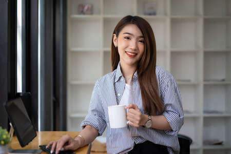 Young beautiful woman holding coffee cup and feeling fresh while sitting at her working place at monday morning.の写真素材