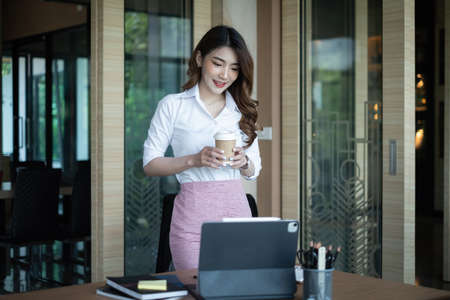 Portrait of cheerful asian woman wear white shirt looking at tablet on desk in office. Concept of young business peopleの写真素材