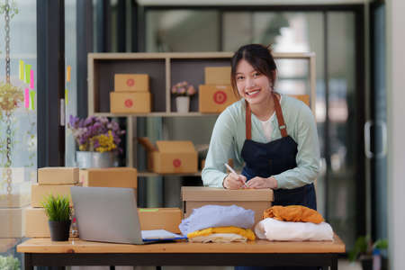 Image of Young asian woman startup freelance with parcel box and talking with customer by computer...