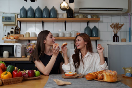 Beautiful woman and friend talking and using laptop at kitchen room. Girl friends couple living...