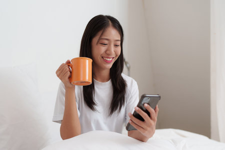 Smiling young asian woman using smart phone in bedroom at home. lifestyle, woman lying on bedの写真素材