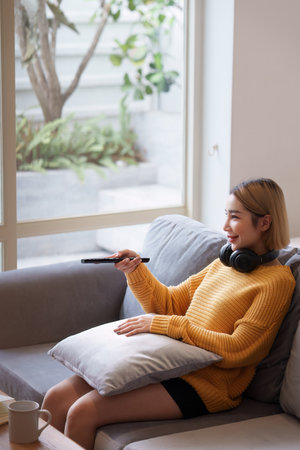 Young woman watching TV resting on sofa sitting in living room in house. Female watch film on television sitting on couch in apartment, leisureの写真素材
