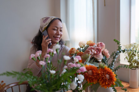 Startup, small business, flower shop. Older florist putting a flower on a vase and talking with customer by phoneの写真素材