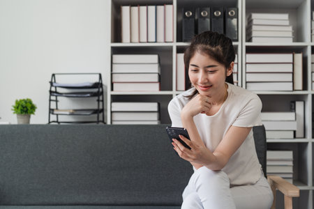 Young relaxed smiling pretty woman relaxing sitting on sofa at home. Female feeling joy enjoying with mobile phone on cozy couchの写真素材