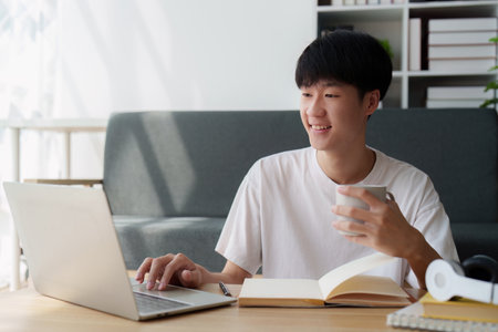 Online education, e-learning. Asian man in stylish casual clothes, studying using a laptop, listening to online lecture, taking notes, online study at homeの写真素材