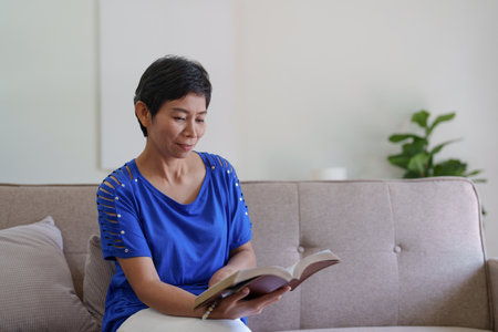 Cheerful asian senior woman reading a book and sitting on sofa at homeの写真素材