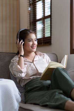 Young relaxed smiling pretty woman relaxing sitting on sofa at home. Female feeling joy enjoying and reading book on cozy couchの写真素材