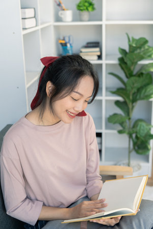 Young Asian woman reading a book at her home sitting on the sofa In the living room
