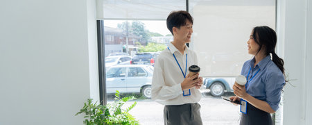 two young asian businesswomen standing chatting talking and holding coffee by the window in office