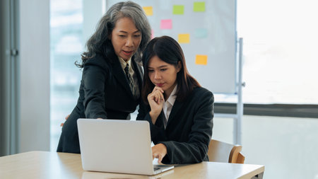 young financial assistant working on laptop while senior professional standing and giving investment adviseの写真素材