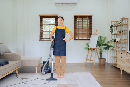 Beautiful girl housewife housekeeper hold vacuum cleaner and smile, looking at camera after doing housework or chores at homeの写真素材