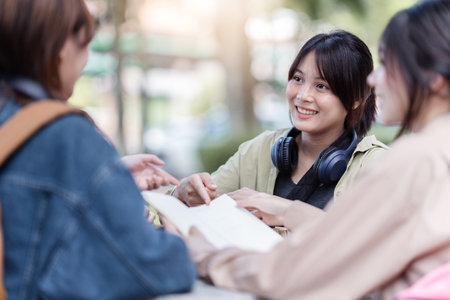 Group of young Asian college students sitting on a bench in a campus relaxation area, talking, sharing ideas, doing homework or tutoring for the exam togetherの写真素材