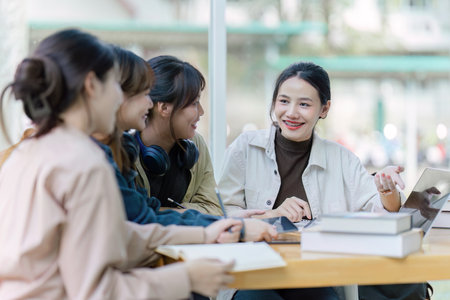 Group of young Asian college students sitting on a bench in a campus relaxation area, talking, sharing ideas, doing homework or tutoring for the exam togetherの写真素材