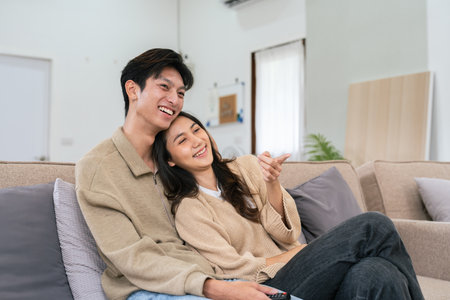Couple Watches TV together while Sitting on a Couch in the Living Room. Girlfriend and Boyfriend embrace, cuddle, talk, smile and watch Television Streaming Servicesの写真素材