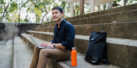 Young businessman working online with laptop while sitting on bench outside of office buildingの写真素材