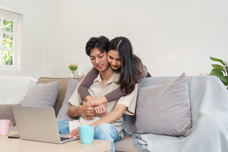 Young Couple Enjoying Quality Time Together at Home, Relaxing on the Couch with a Laptop, Smiling and Embracing in a Cozy Living Room Setting, Perfect for Lifestyle and Home Comfort Themesの写真素材