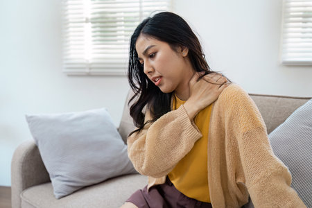 Woman Experiencing Shoulder Pain While Sitting on Sofa at Home, Holding Neck in Discomfortの写真素材