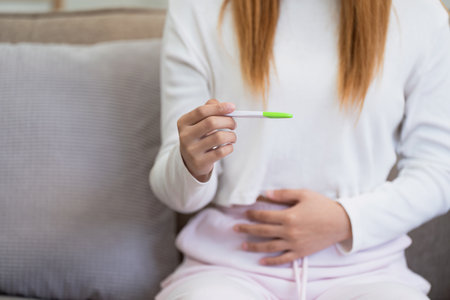 Woman Holding Positive Pregnancy Test While Touching Her Stomach, Celebrating Early Pregnancy at Homeの写真素材