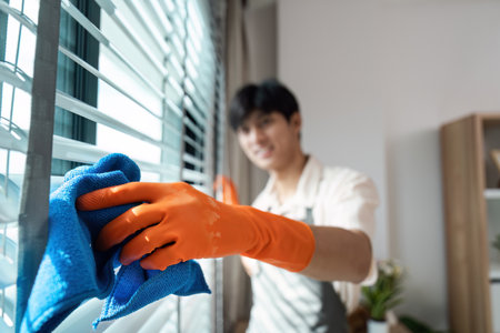 Young Man Cleaning Window Blinds with Blue Cloth and Orange Gloves in Modern Home Interior