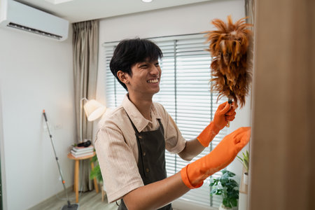 Young Man Smiling While Dusting Furniture in Modern Living Room with Orange Gloves and Feather...