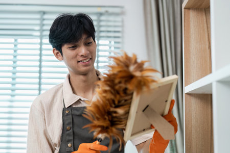 Young Man Dusting Picture Frame with Feather Duster in Modern Home, Wearing Apron and Gloves, Bright...