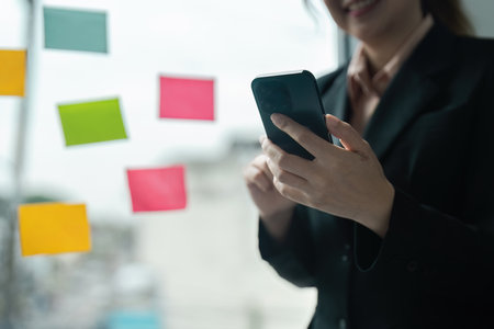 Professional Businesswoman Using Smartphone in Modern Office with Colorful Sticky Notes on Glass Wallの写真素材