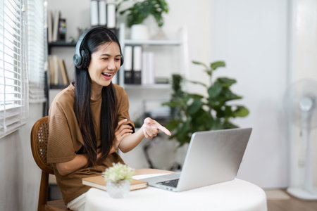 Young Woman Using Laptop with Headphones in Modern Home Office Setting, Engaging in Virtual Meeting...