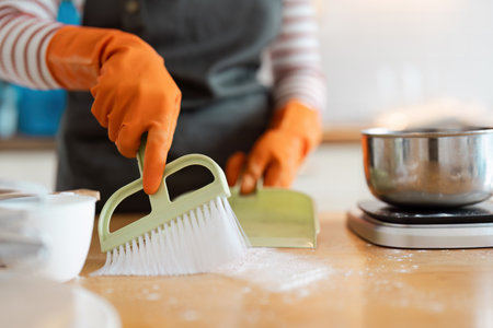 Woman Cleaning Kitchen Counter with Dustpan and Brush Wearing Orange Gloves in Modern Home