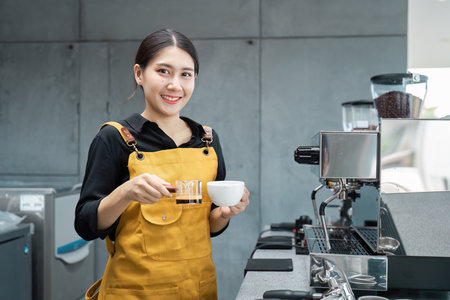 Young Female Barista in Yellow Apron Holding Coffee Cup and Smiling in Modern Cafeの写真素材