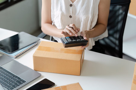 Small Business Owner Calculating Costs for Online Sales with Laptop and Packaging Box on Deskの写真素材