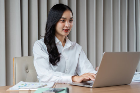 Young Professional Woman Working on Laptop in Modern Office Setting with Documents and Chartsの写真素材