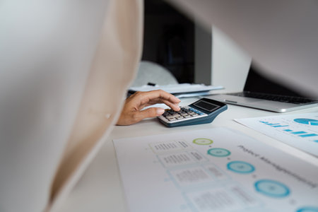 Business Professional Using Calculator for Financial Analysis at Modern Office Desk with Graphs and Laptopの写真素材