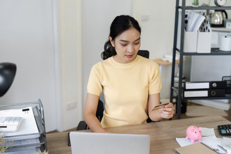 Young Woman Engaged in Financial Planning and Savings Management at Home Office with Laptop, Credit Card, and Piggy Bankの写真素材