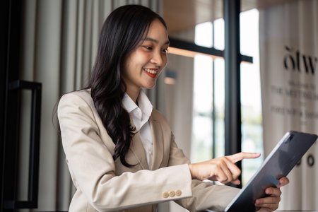 Businesswoman in Modern Office Using Tablet for Working, Smiling and Engaged in Digital Communication, Professional Attire, Bright and Contemporary Settingの写真素材