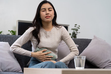 Young Woman Experiencing Stomach Pain While Sitting on a Couch Holding a Hot Water Bottle for Relief in a Cozy Living Room Settingの写真素材