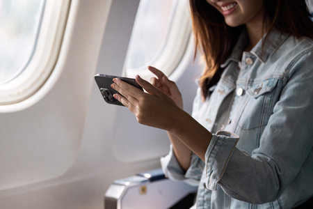 Woman Enjoying In-Flight Entertainment on Smartphone While Traveling on Airplane with Sunlight Streaming Through Windowの写真素材