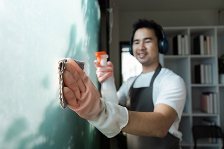 Smiling Man Cleaning Glass with Spray Bottle and Cloth in Modern Home Environmentの写真素材