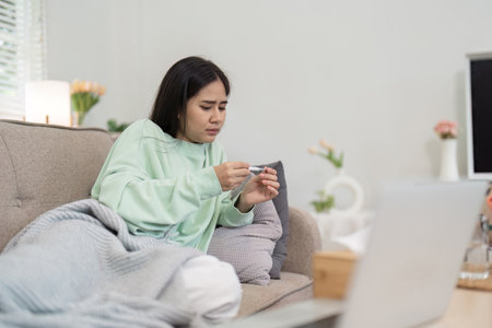 Woman Checking Temperature at Home While Feeling Unwell, Wrapped in Blanket on Sofa, Illustrating...