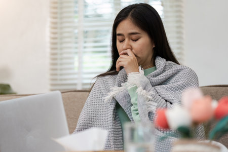 Woman Feeling Unwell at Home Wrapped in Blanket with Laptop and Tissues Nearby