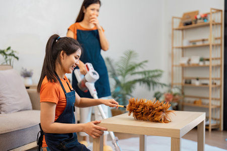 Daughter dusting the table while mother supervises cleaning effortsの写真素材