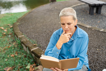 Thoughtful senior woman pondering while reading notes from her notebook by the lakeside in a park