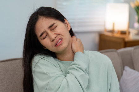 Woman in a mint green sweater grimacing in pain at home, clutching her neck to relieve discomfort and express distressの写真素材